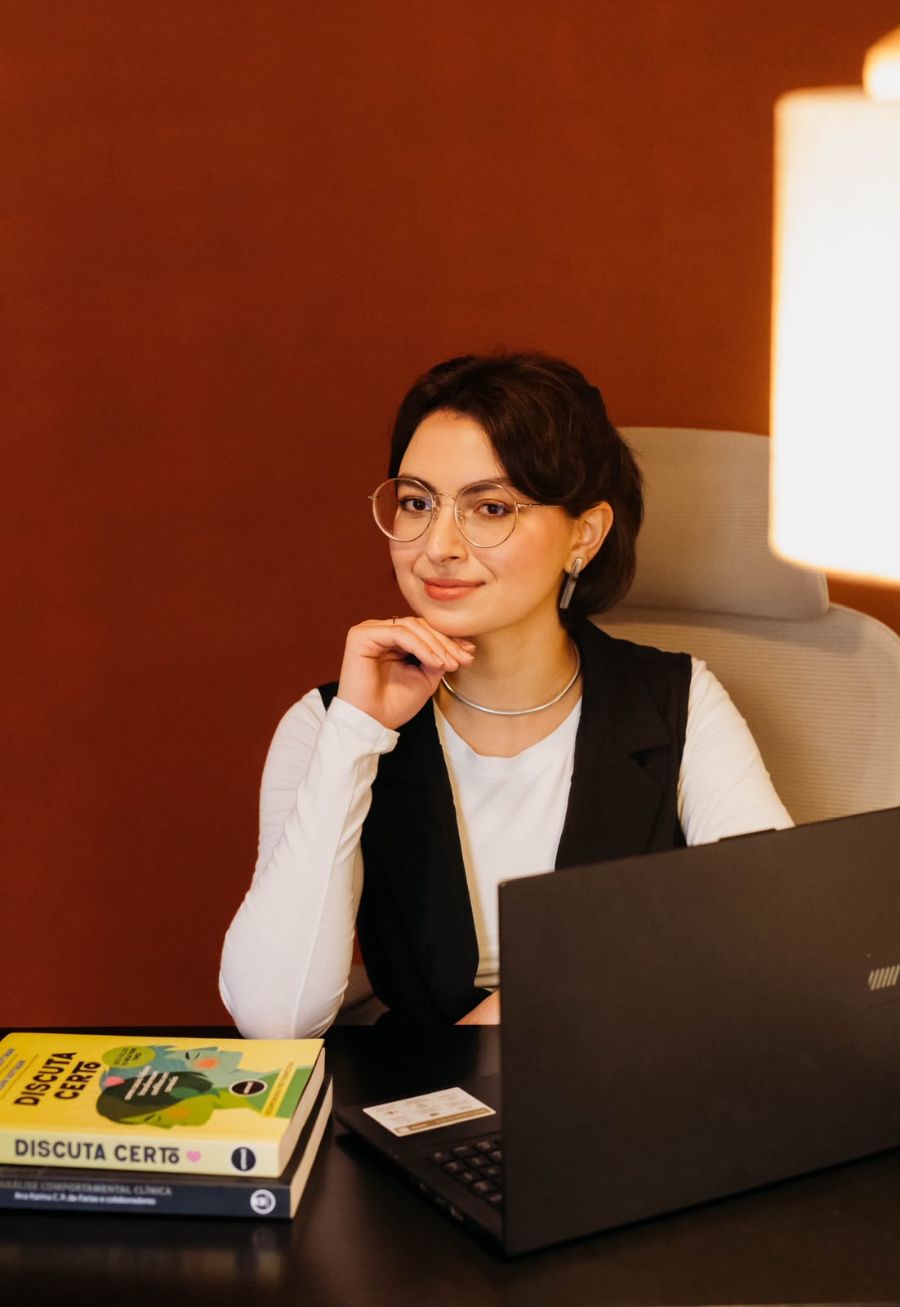 Jade Teixeira à mesa do consultório, com a mão no queixo, livros de análise comportamental ao lado